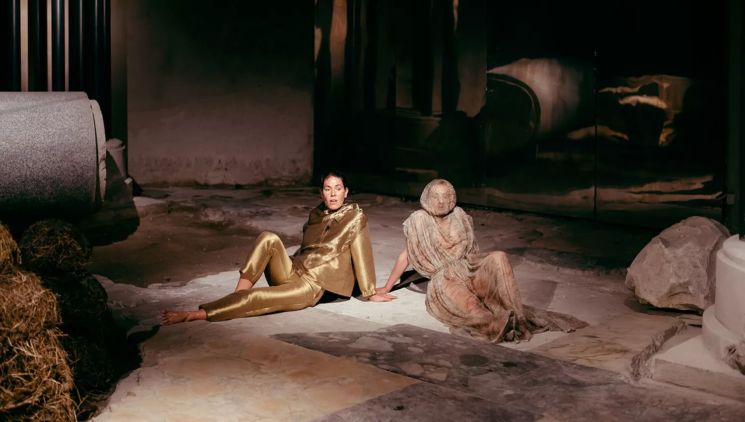 Gaia Fugazza - Gaia Fugazza, 2024, Farewell to Known Skies. Two performers sit at the centre of a beige tiled floor: one wears a gold suit, while the other is covered in netting. A dark, indistinct background surrounds them.