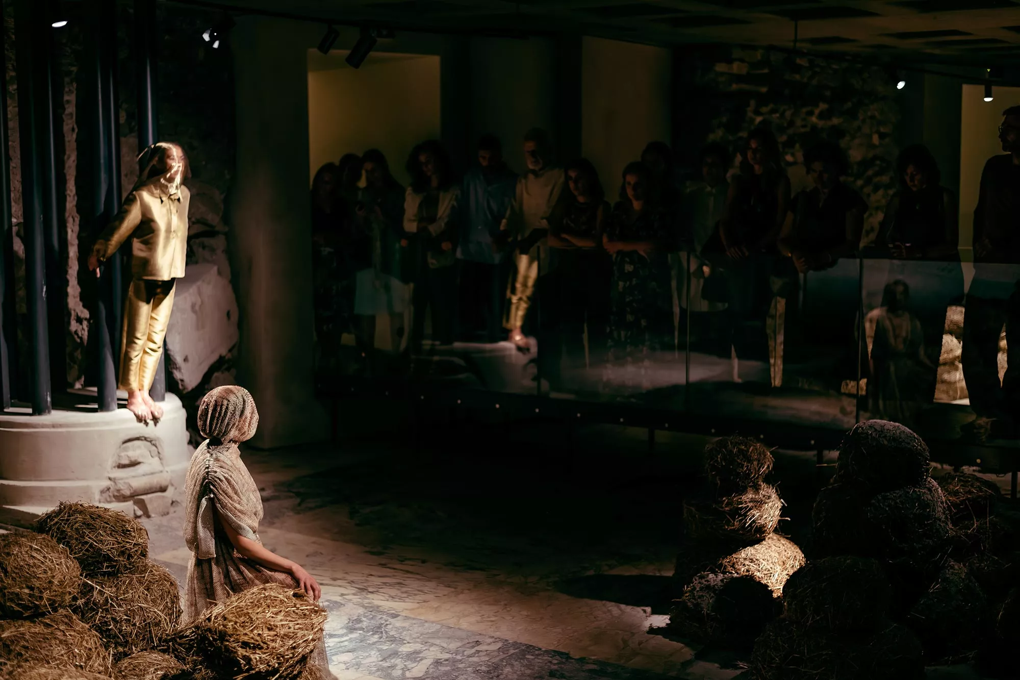 Gaia Fugazza - Gaia Fugazza, 2024, Fairwell to Known Skies. Audience-facing view of the performance, two performers stand on the left side in a dark, atomospheric setting.