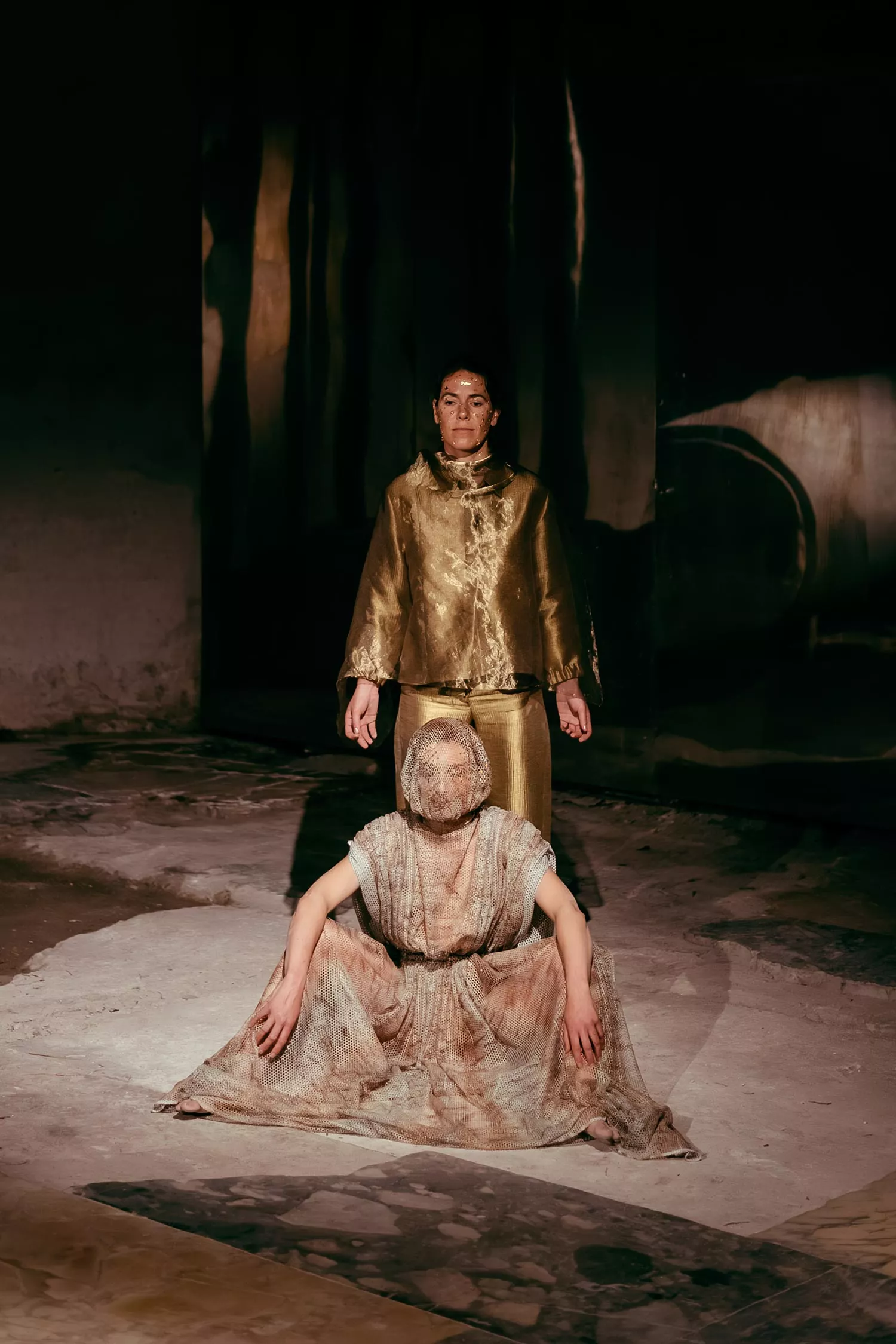 Gaia Fugazza - Gaia Fugazza, 2024, Farewell to Known Skies. Two performers are positioned at the centre of a shadowy, stone-filled setting, with one standing behind the other, who sits in front.