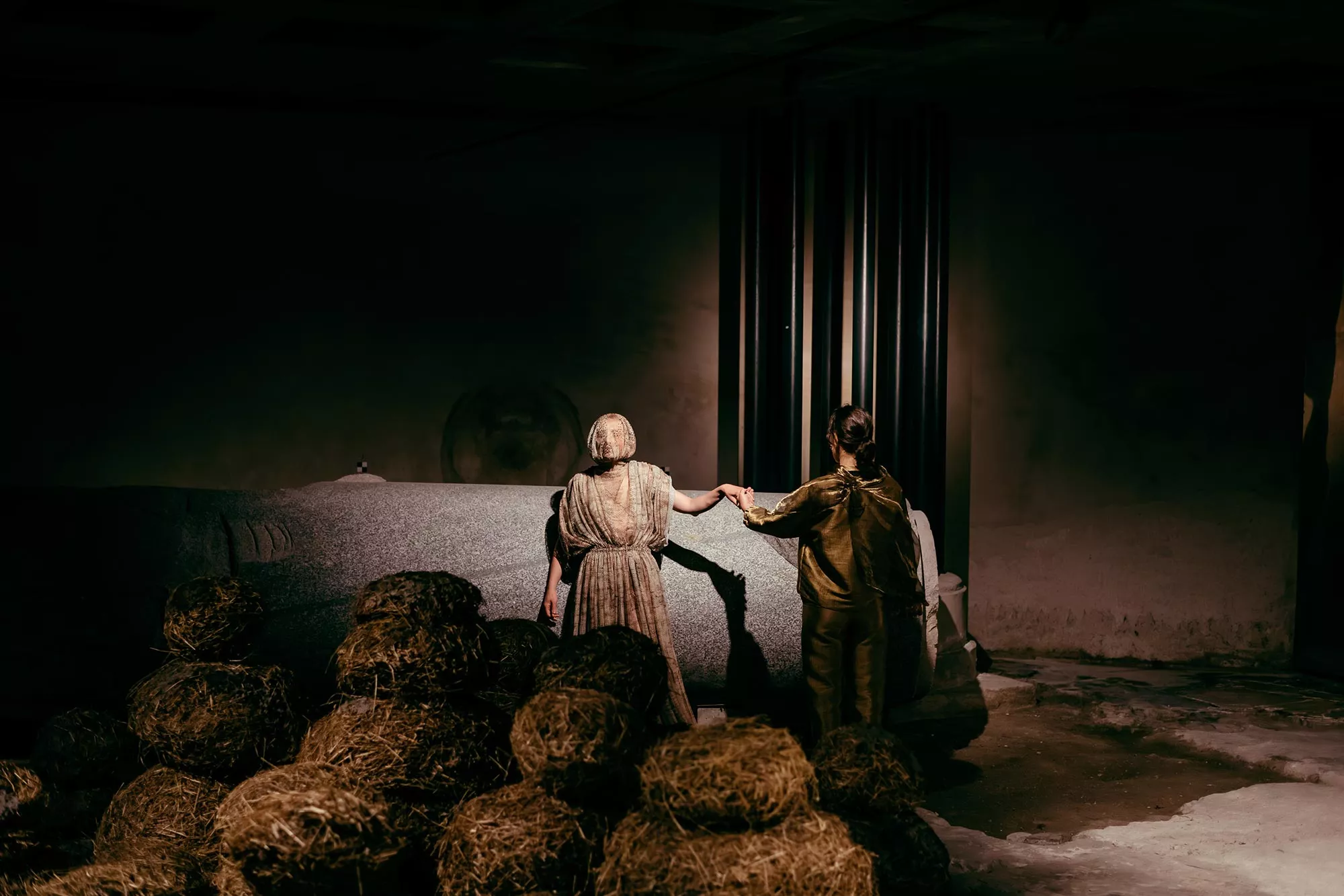 Gaia Fugazza - Gaia Fugazza, 2024, Farewell to Known Skies. One performer takes another's hand, behind a hay installation in the foreground. They are set against a dark, shadowy background, with a bare stone floor beneath them.