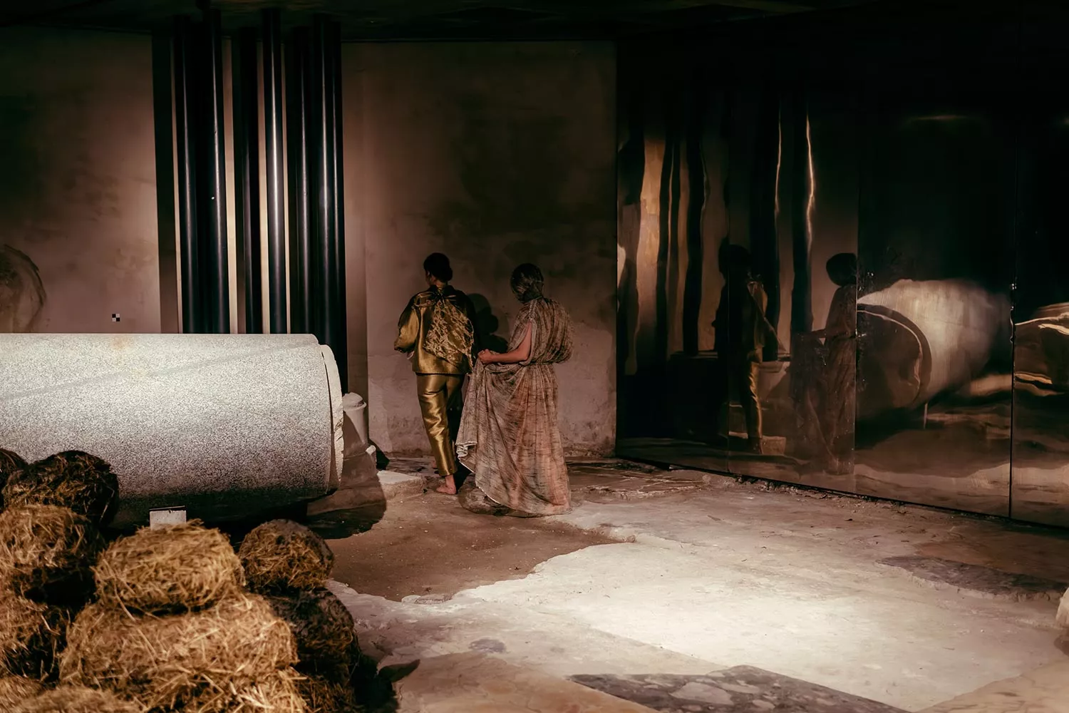 Gaia Fugazza - Gaia Fugazza, 2024, Farewell to Known Skies. Two performers walk away side by side with their backs to the audience, beside a mirror. Hay structures stand to the left, and a bare stone floor fills the space.